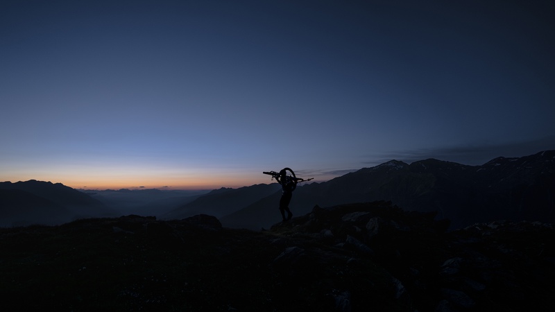 Young mountain bike athlete Bo Schwarz carries a mountain bike on his back. Wearing a bright headlamp he is silhouetted with the smooth morning colors of blue and orange filling the mountain landscape behind him.