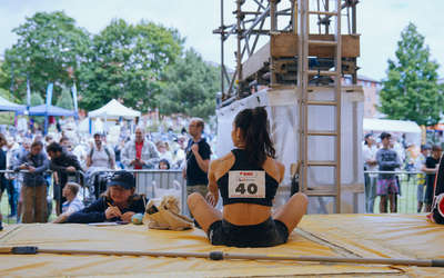 A woman with dark hair and a competitors sticker on her top sits with her back to us on climbing matts looking out over a large crowd.