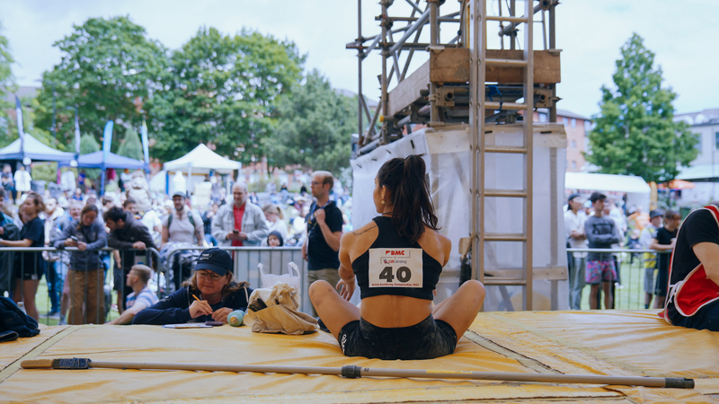 A woman with dark hair and a competitors sticker on her top sits with her back to us on climbing matts looking out over a large crowd.