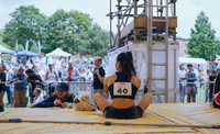 A woman with dark hair and a competitors sticker on her top sits with her back to us on climbing matts looking out over a large crowd.