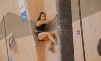 A woman with dark hair has pinned herself between the climbing wall and a huge volume climbing hold, facing us as she goes up the chimney her tongue is poking out with effort!