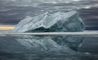 Expansive Arctic scene featuring a massive iceberg reflected perfectly on the calm, glass-like surface of the water beneath a moody, overcast sky. The iceberg’s textured, rugged surface contrasts with the smoothness of the reflection, creating a mirror image that enhances the sense of solitude and vastness in the Arctic landscape. The muted light and layered clouds evoke a serene yet somber atmosphere, highlighting the beauty and vulnerability of this icy giant. 