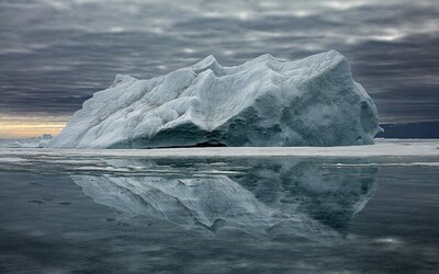 Expansive Arctic scene featuring a massive iceberg reflected perfectly on the calm, glass-like surface of the water beneath a moody, overcast sky. The iceberg’s textured, rugged surface contrasts with the smoothness of the reflection, creating a mirror image that enhances the sense of solitude and vastness in the Arctic landscape. The muted light and layered clouds evoke a serene yet somber atmosphere, highlighting the beauty and vulnerability of this icy giant. 
