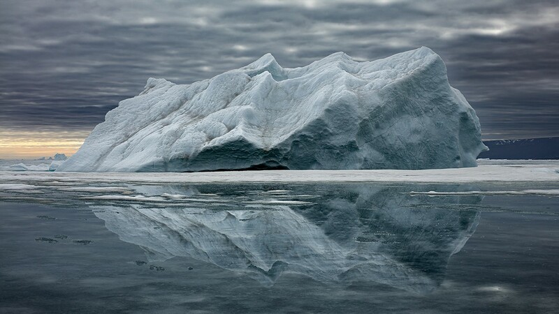 Expansive Arctic scene featuring a massive iceberg reflected perfectly on the calm, glass-like surface of the water beneath a moody, overcast sky. The iceberg’s textured, rugged surface contrasts with the smoothness of the reflection, creating a mirror image that enhances the sense of solitude and vastness in the Arctic landscape. The muted light and layered clouds evoke a serene yet somber atmosphere, highlighting the beauty and vulnerability of this icy giant. 