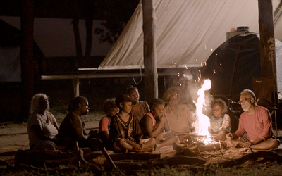 In this photo, a group of people of various ages sit closely around a campfire at night, illuminated by its warm glow. Dean is the most visibly lit by the fire’s light. The setting is outdoors under a large tarp, creating an intimate and communal atmosphere.