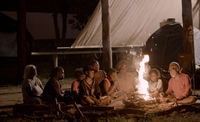 In this photo, a group of people of various ages sit closely around a campfire at night, illuminated by its warm glow. Dean is the most visibly lit by the fire’s light. The setting is outdoors under a large tarp, creating an intimate and communal atmosphere.