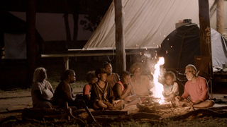 In this photo, a group of people of various ages sit closely around a campfire at night, illuminated by its warm glow. Dean is the most visibly lit by the fire’s light. The setting is outdoors under a large tarp, creating an intimate and communal atmosphere.