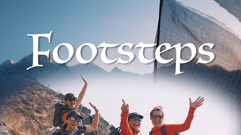 The four female members of the Footsteps expedition team stand and wave on the side of a mountain. They are wearing expedition rucksacks and himalayan peaks can be seen in the background. Text 'Footsteps' is written across the image with the JHPVisuals, Rab, La Sportiva and Exped brand logos also visible.