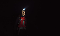 In vast darkness, the upper body of a woman can be seen. She is wearing a head torch, the light of which illuminates the frost of her breath. She is wearing a red soft-shell, with a rucksack over her shoulders, strap tightened across her chest. She stares into the darkness, to the side of the camera, in contemplation and curiosity.