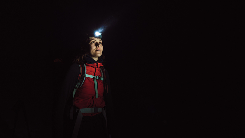 In vast darkness, the upper body of a woman can be seen. She is wearing a head torch, the light of which illuminates the frost of her breath. She is wearing a red soft-shell, with a rucksack over her shoulders, strap tightened across her chest. She stares into the darkness, to the side of the camera, in contemplation and curiosity.