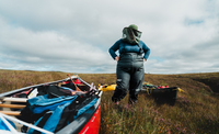 Under a cloudy sky, a plus-sized woman in a dark blue thermal and dark grey wading trousers stands with her hands on her hips. She is wearing a hat & midge net that obscures her face. By her feet, a bright red canoe laden with luggage fills the lower left of the image, going out of frame. Surrounding her, and the canoe, is thick heathery bog. 