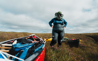 Under a cloudy sky, a plus-sized woman in a dark blue thermal and dark grey wading trousers stands with her hands on her hips. She is wearing a hat & midge net that obscures her face. By her feet, a bright red canoe laden with luggage fills the lower left of the image, going out of frame. Surrounding her, and the canoe, is thick heathery bog. 