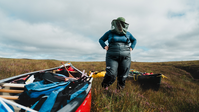 Under a cloudy sky, a plus-sized woman in a dark blue thermal and dark grey wading trousers stands with her hands on her hips. She is wearing a hat & midge net that obscures her face. By her feet, a bright red canoe laden with luggage fills the lower left of the image, going out of frame. Surrounding her, and the canoe, is thick heathery bog. 