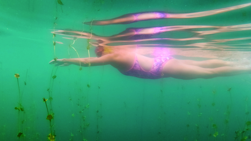 Melanie, a white woman in a brighly patterned swimsuit glides under the surface of a lake, her arms stretched out front, legs gently kicking up bubbles behind. Leafy tendrils rise up from the bottom, surrounding her body, which is reflected on the underside of the lake above her.