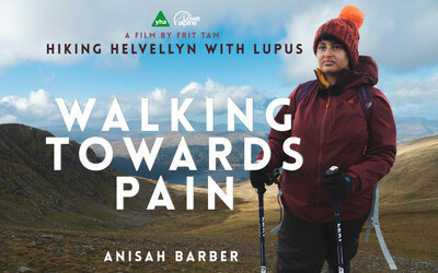 A young south asian woman dressed in a bobble hat, waterproof jacket and gloves, holds hiking poles and stares into the distance. The words "Walking Towards Pain" are large, alongside "Hiking Helvellyn with Lupus" and "Anisah Barber" - the main character in the film