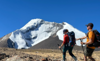 A man follows a woman by touching their backpack. They both hold walking poles. A snowy mountain can be seen behind them.