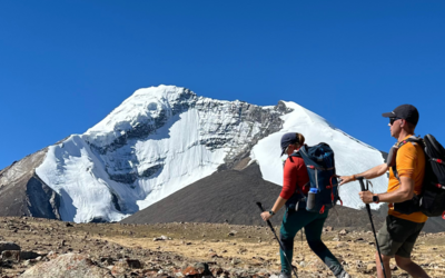 A man follows a woman by touching their backpack. They both hold walking poles. A snowy mountain can be seen behind them.