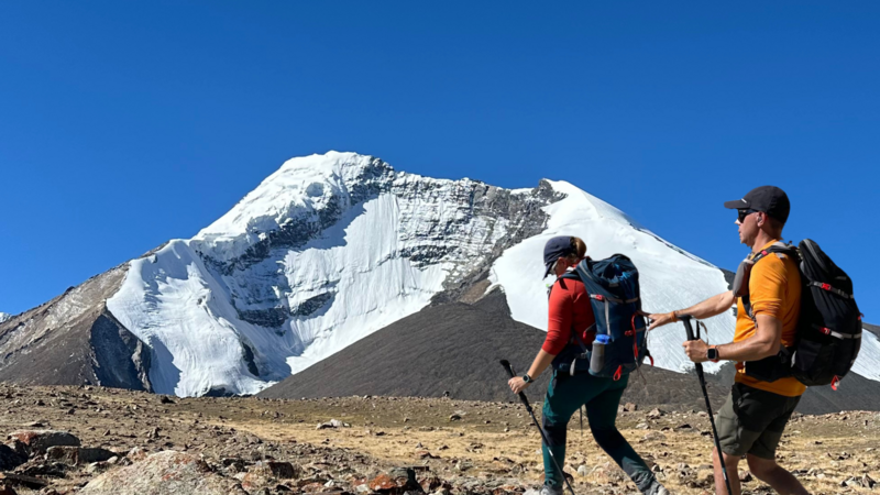 A man follows a woman by touching their backpack. They both hold walking poles. A snowy mountain can be seen behind them.