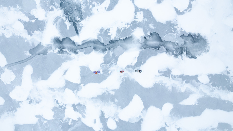 looking top down on skiers crossing a frozen loch with patchy wind blown snow covering the ice