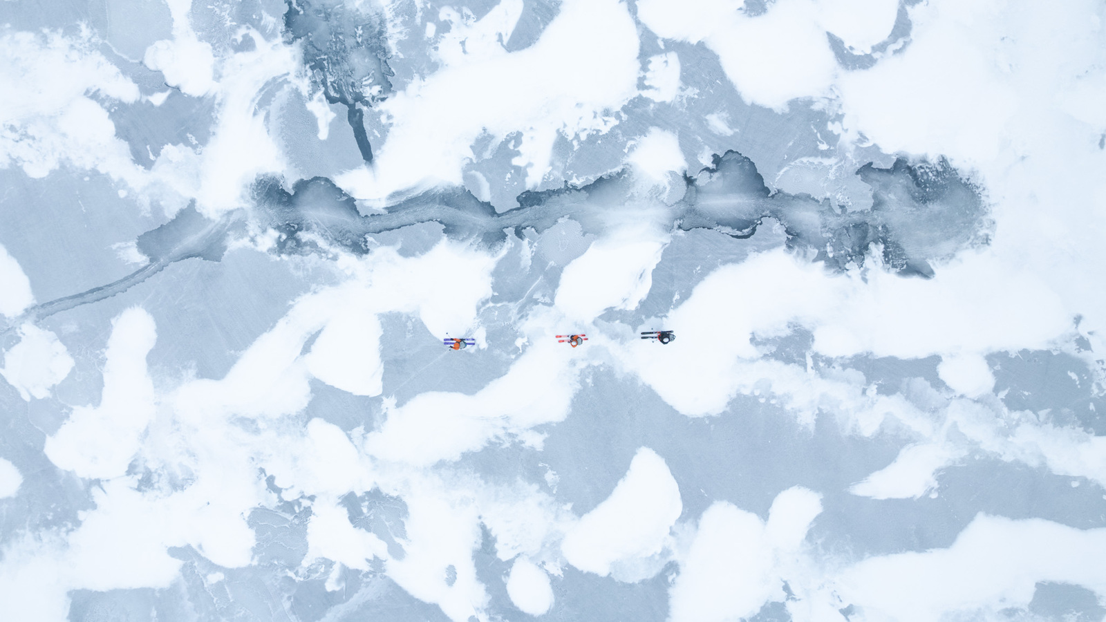 looking top down on skiers crossing a frozen loch with patchy wind blown snow covering the ice