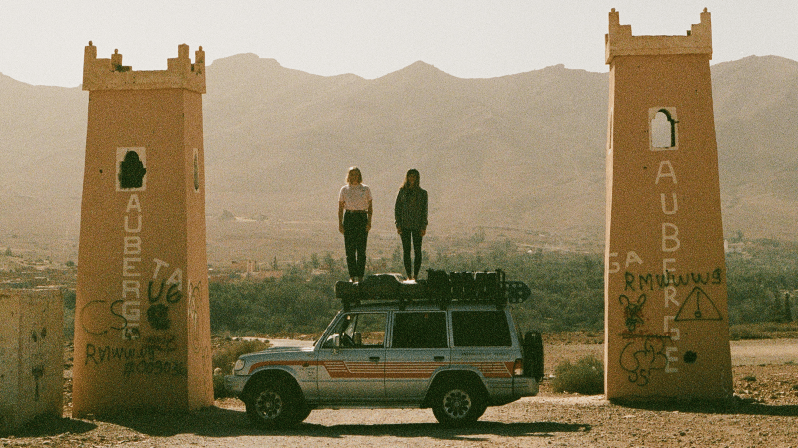 Two women stand on the roof of a loaded 4x4 vehicle parked between two tall, weathered gateway towers in a desert landscape, with distant mountains in the background.