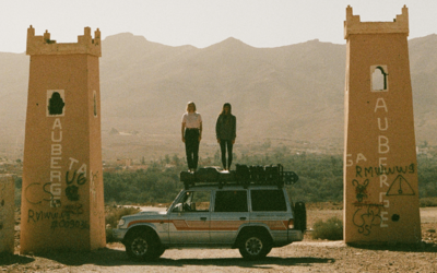 Two women stand on the roof of a loaded 4x4 vehicle parked between two tall, weathered gateway towers in a desert landscape, with distant mountains in the background.