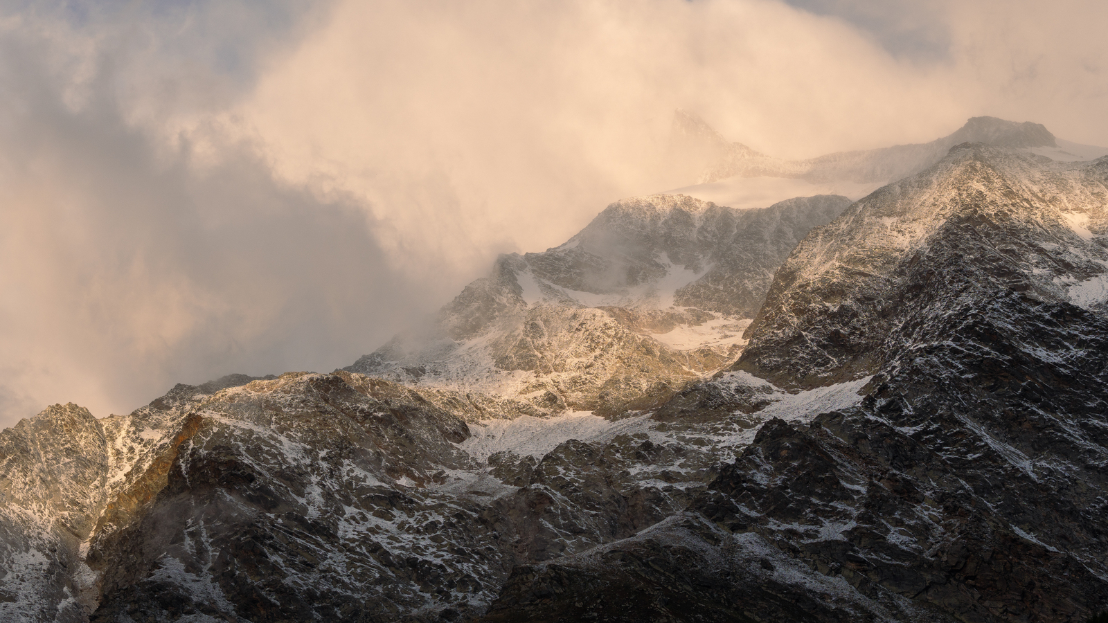 Sunlight breaks through clouds to illuminate a rugged mountain range partially covered with snow, creating a dramatic contrast between the warm glow on the peaks and the dark, shadowed slopes below.
