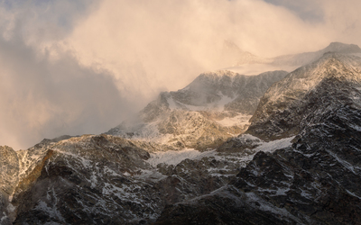 Sunlight breaks through clouds to illuminate a rugged mountain range partially covered with snow, creating a dramatic contrast between the warm glow on the peaks and the dark, shadowed slopes below.