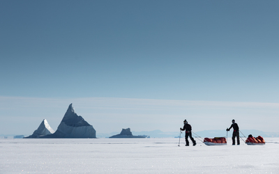 Bright arctic scene with two split boarders pulling pulks in front of distant icebergs frozen into the ice. One is in motion, the other is standing still.
