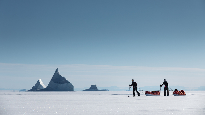 Bright arctic scene with two split boarders pulling pulks in front of distant icebergs frozen into the ice. One is in motion, the other is standing still.