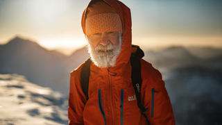 A close-up portrait of Nick Gardner, on a snowy summit, ice in his beard, and with the sun low in the sky behind him.