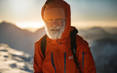 A close-up portrait of Nick Gardner, on a snowy summit, ice in his beard, and with the sun low in the sky behind him. 