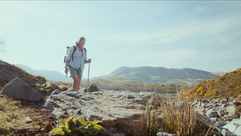 A lady is holding a stick and using it to support her as she slowly walks up the fell side with the sun shining on her back. Her face is exasperated.
