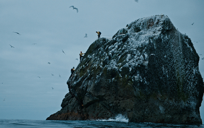 Three people climb up a small rock, surrounded by ocean.