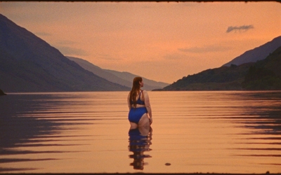 Swimmer stands in water up to her thighs in a loch at sunset surrounded by mountains
