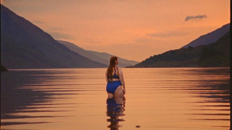 Swimmer stands in water up to her thighs in a loch at sunset surrounded by mountains