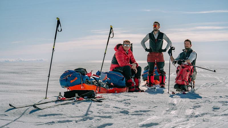 Three men, one sat on a pulk, one stood, and one sat in a specialised wheelchair on top of an icecap. 