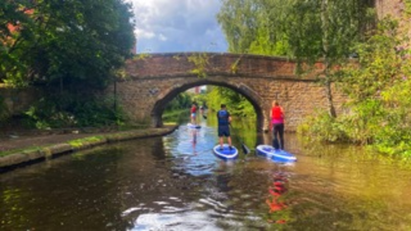 Three people stand on Stand up paddle boards approaching a bridge on a Canal