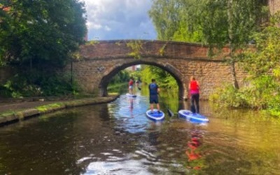 Three people stand on Stand up paddle boards approaching a bridge on a Canal